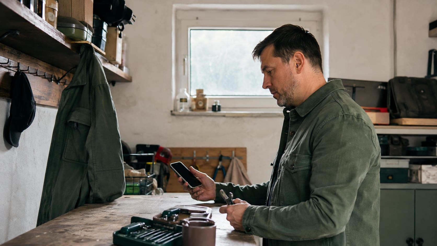 Mężczyzna stoi przy warsztatowym blacie i trzyma telefon z pustym ekranem oraz narzędzie Mężczyzna stoi przy warsztatowym blacie i trzyma telefon z pustym ekranem oraz narzędzie